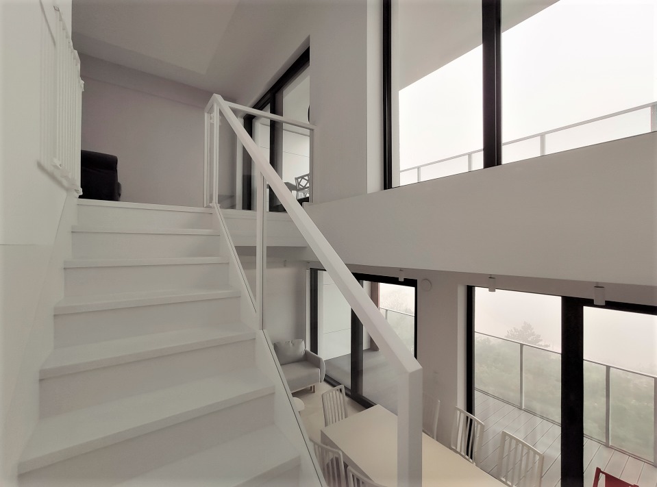 Minimalistyczne wnętrze z białymi schodami z przeszkloną balustradą, widok na duży salon z oknami panoramicznymi i stołem z krzesłami, nowoczesny design.