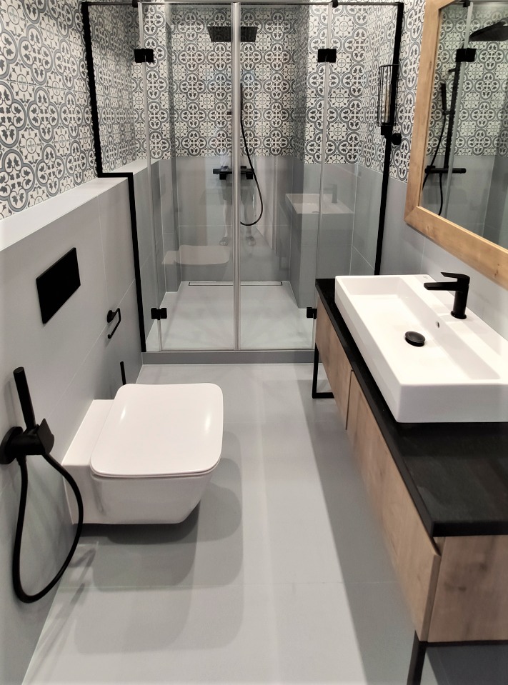 Nowoczesna łazienka w Krakowie po remoncie, z wiszącą toaletą z czarną armaturą, kabiną prysznicową w czarnych ramach i umywalką nablatową na szafce z drewnopodobnym frontem.