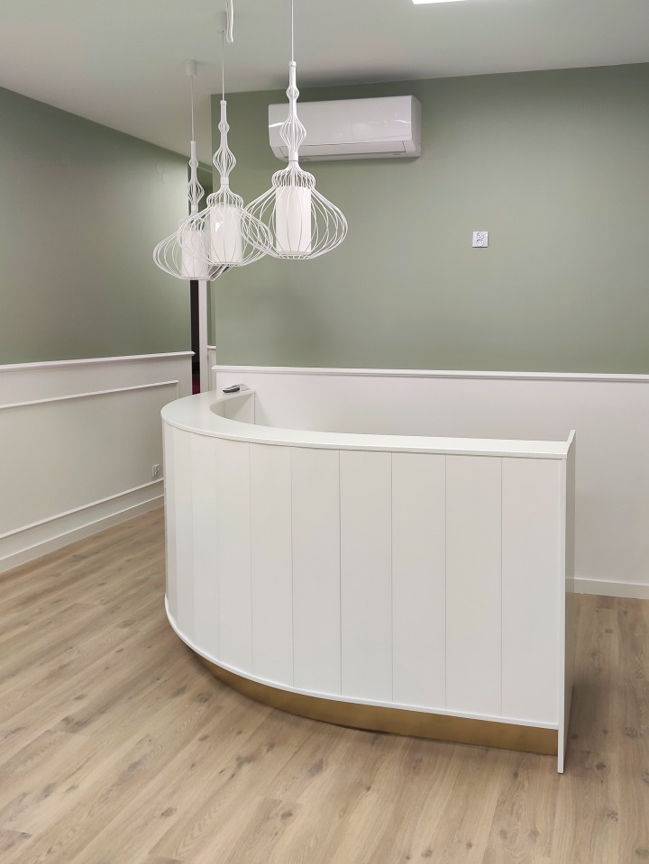 Nowoczesne wnętrze biura z zaokrągloną białą recepcją z złotą podstawą, trzema wiszącymi lampami z białymi abażurami i klimatyzacją na pistacjowej ścianie.