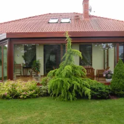 Zadaszony taras z brązowymi ramami w Dębnie, widok z zadbanego ogrodu, z roślinami ozdobnymi i meblami ogrodowymi wewnątrz.