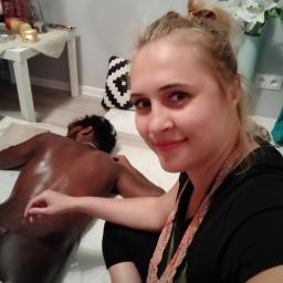 Masaż relaksacyjny - Kobieta uśmiecha się do kamery podczas wykonywania masażu pleców u mężczyzny na białym prześcieradle, w tle stolik z dekoracjami i kwiaty.