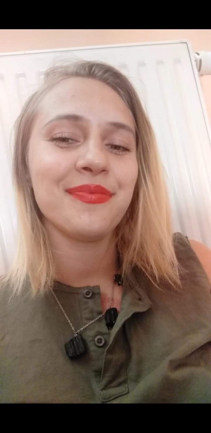 Portret kobiety z blond włosami i intensywnie czerwoną pomadką, ubrana w oliwkową koszulę z naszyjnikiem z czarnych kamieni, na tle białego grzejnika.