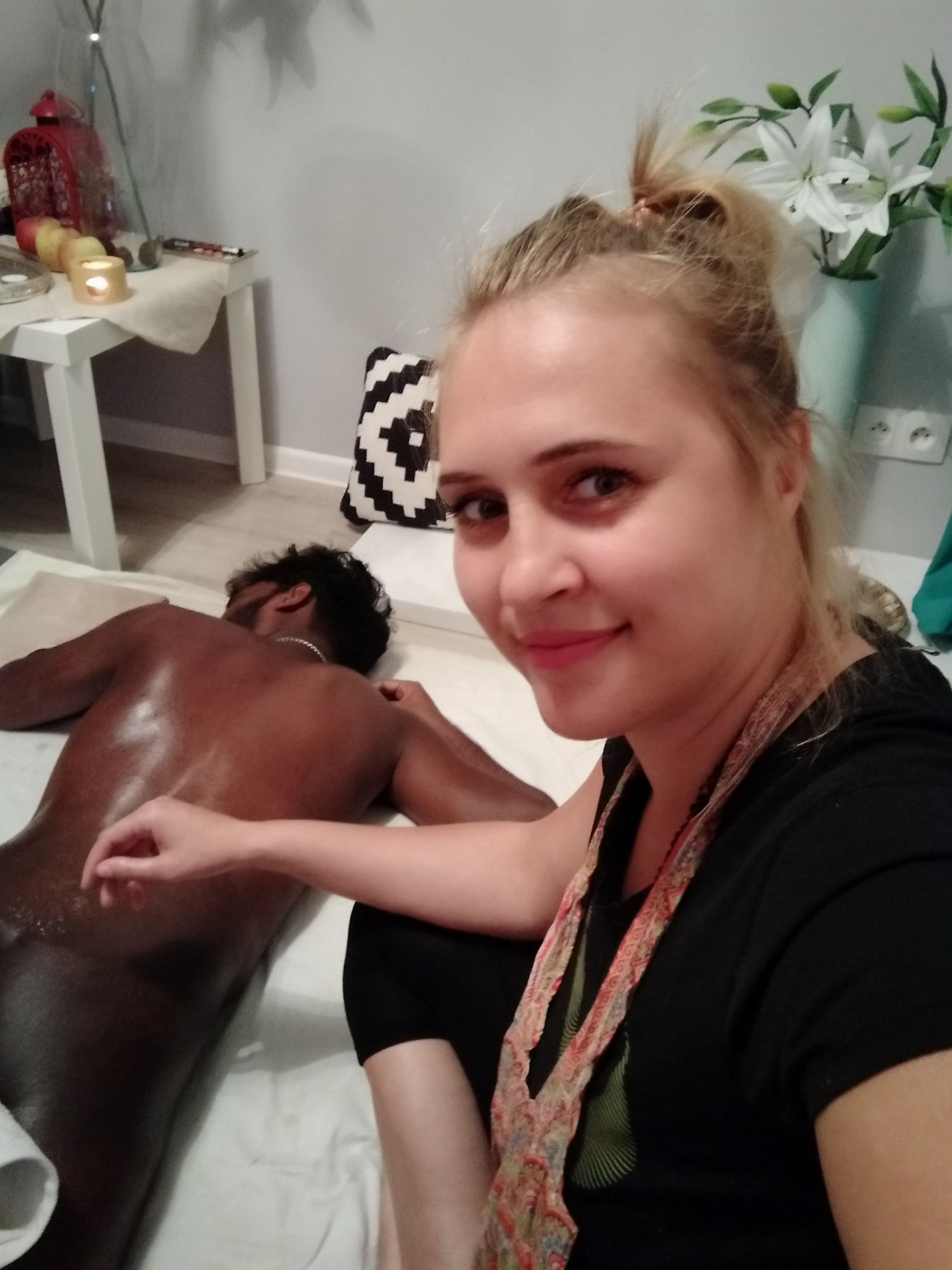 Kobieta uśmiecha się do kamery podczas wykonywania masażu pleców u mężczyzny na białym prześcieradle, w tle stolik z dekoracjami i kwiaty.