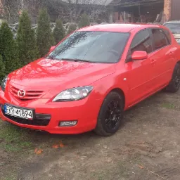 Czerwona Mazda 3 hatchback z czarnymi felgami stoi na trawiasto-błotnistym podłożu przed garażem i rzędem tuj. Na pierwszym planie widoczna tablica rejestracyjna z polską flagą.