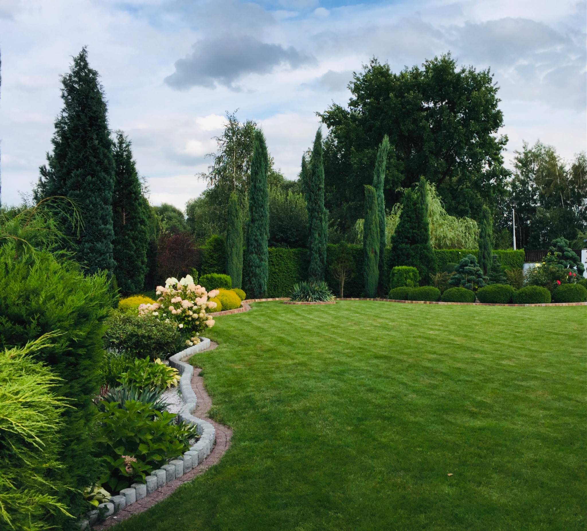 Zadbany ogród z równo przystrzyżonym trawnikiem, ozdobnymi rabatami kwiatowymi z obrzeżami z kostki i różnorodnymi drzewami iglastymi oraz liściastymi w tle.