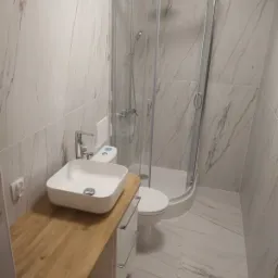 Nowoczesna łazienka z kabiną prysznicową, umywalką nablatową na drewnianym blacie i marmurowymi płytkami na ścianach oraz podłodze.