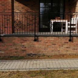 VIZIA Piotr Jankowiak - Czarna, metalowa balustrada balkonowa z ozdobnymi elementami na tle ceglanej ściany domu i brukowanego chodnika.