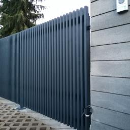 VIZIA Piotr Jankowiak - Nowoczesny płot z profili aluminiowych w kolorze grafitowym, widok z zewnątrz na posesję z kostką ażurową, obok szary mur z dzwonkiem.