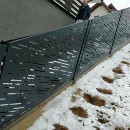 VIZIA Piotr Jankowiak - Nowoczesne ogrodzenie z blachy z poziomymi, prostokątnymi wycięciami, zamontowane na pochyłym terenie pokrytym śniegiem, z widocznymi śladami stóp.