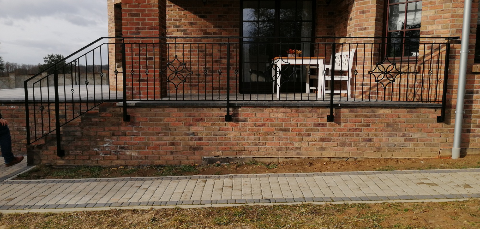 Czarna, metalowa balustrada balkonowa z ozdobnymi elementami na tle ceglanej ściany domu i brukowanego chodnika.