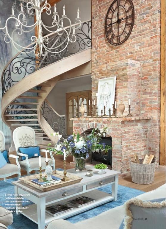 Wnętrze salonu z spiralnymi schodami z drewnianymi stopniami i kutą, ozdobną balustradą, ceglany kominek z dekoracjami, jasne meble i żyrandol w stylu retro.