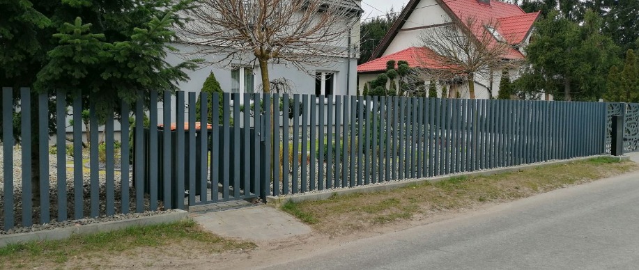 Nowoczesne, grafitowe ogrodzenie posesji z pionowych, wąskich profili, z furtką i ozdobnym panelem ażurowym po prawej stronie, na tle domów i zieleni.