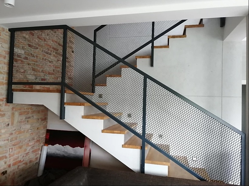 Schody drewniane z metalową, siatkową barierką w stylu loftowym; fragment ściany z cegły w tle.