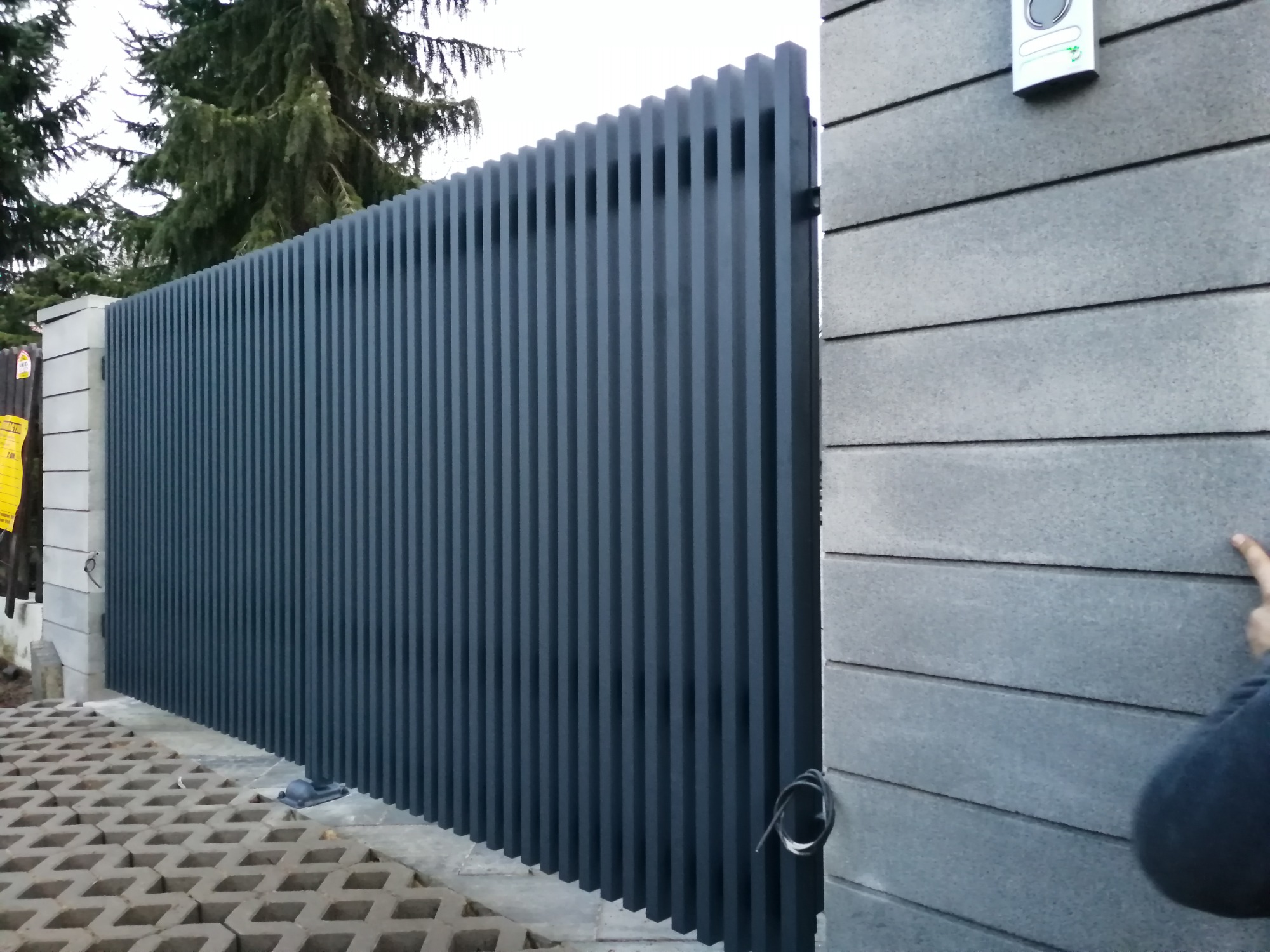 Nowoczesny płot z profili aluminiowych w kolorze grafitowym, widok z zewnątrz na posesję z kostką ażurową, obok szary mur z dzwonkiem.