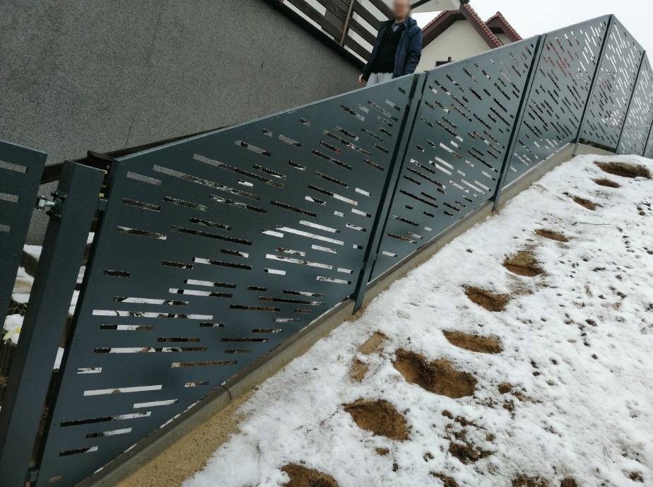 Nowoczesne ogrodzenie z blachy z poziomymi, prostokątnymi wycięciami, zamontowane na pochyłym terenie pokrytym śniegiem, z widocznymi śladami stóp.