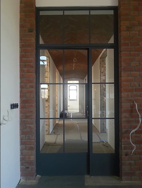 Loftowe drzwi przeszklone z czarną ramą, widok na długi korytarz w trakcie remontu, surowe ceglane ściany po bokach otworu drzwiowego, widoczne przewody elektryczne na ścianie.
