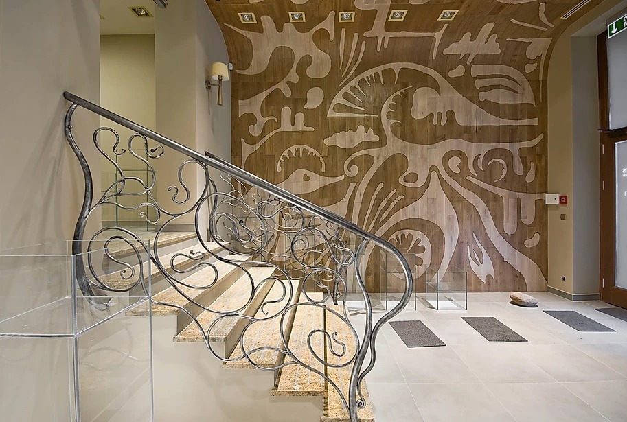 Kuta balustrada schodowa z metalu o dekoracyjnych, roślinnych motywach, połączona z panelami szklanymi, na tle ściany z abstrakcyjnym wzorem w odcieniach beżu.
