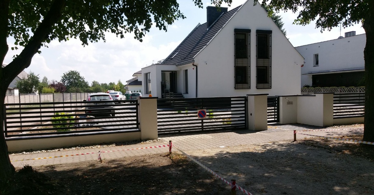 Nowoczesne ogrodzenie posesji z poziomych profili metalowych w kolorze czarnym, z bramą wjazdową i furtką, na tle białego domu z ciemną dachówką.
