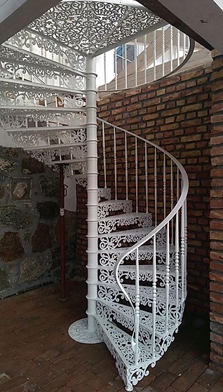 Białe, metalowe schody spiralne z dekoracyjnymi, ażurowymi elementami, wkomponowane w rustykalne wnętrze z ceglaną ścianą i kamienną posadzką.