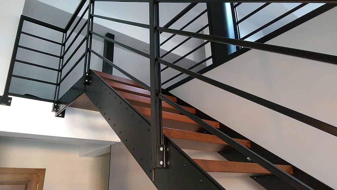 Schody loftowe z drewnianymi stopniami i czarną, metalową balustradą o minimalistycznym designie, mocowaną na śruby.