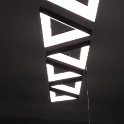 Geometryczna lampa sufitowa z trójkątnych modułów LED, zawieszona na cienkich linkach, z widocznym kablem zasilającym i oknem w tle.