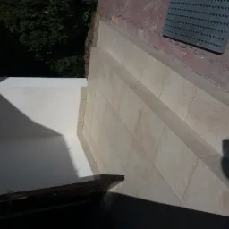 Wyłożone jasnymi płytkami schody wejściowe do budynku, widoczny fragment ściany i cień osoby robiącej zdjęcie.