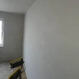 Świeżo wykończona, gładka ściana w szarym odcieniu, oświetlona dwoma przenośnymi lampami roboczymi z żółtymi elementami, widoczny fragment okna i surowa podłoga w tle.