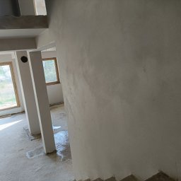 PAW-BUD - Ściana po nałożeniu tynku maszynowego w surowym wnętrzu budynku z oknami. Widoczne ślady wody na posadzce i kolumny konstrukcyjne.