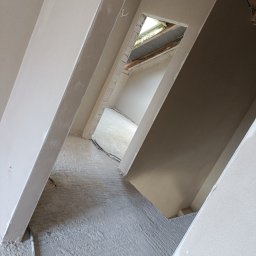 PAW-BUD - Wnętrze po tynkowaniu maszynowym: świeżo otynkowane ściany i podłoga, widoczne kable, surowy stan wykończenia z oknem w dachu w tle, perspektywa z dołu.