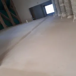 Świeżo wylany, jasny tynk maszynowy na podłodze w surowym wnętrzu, widoczne drewniane elementy konstrukcyjne dachu i surowe ściany z oknem w tle.