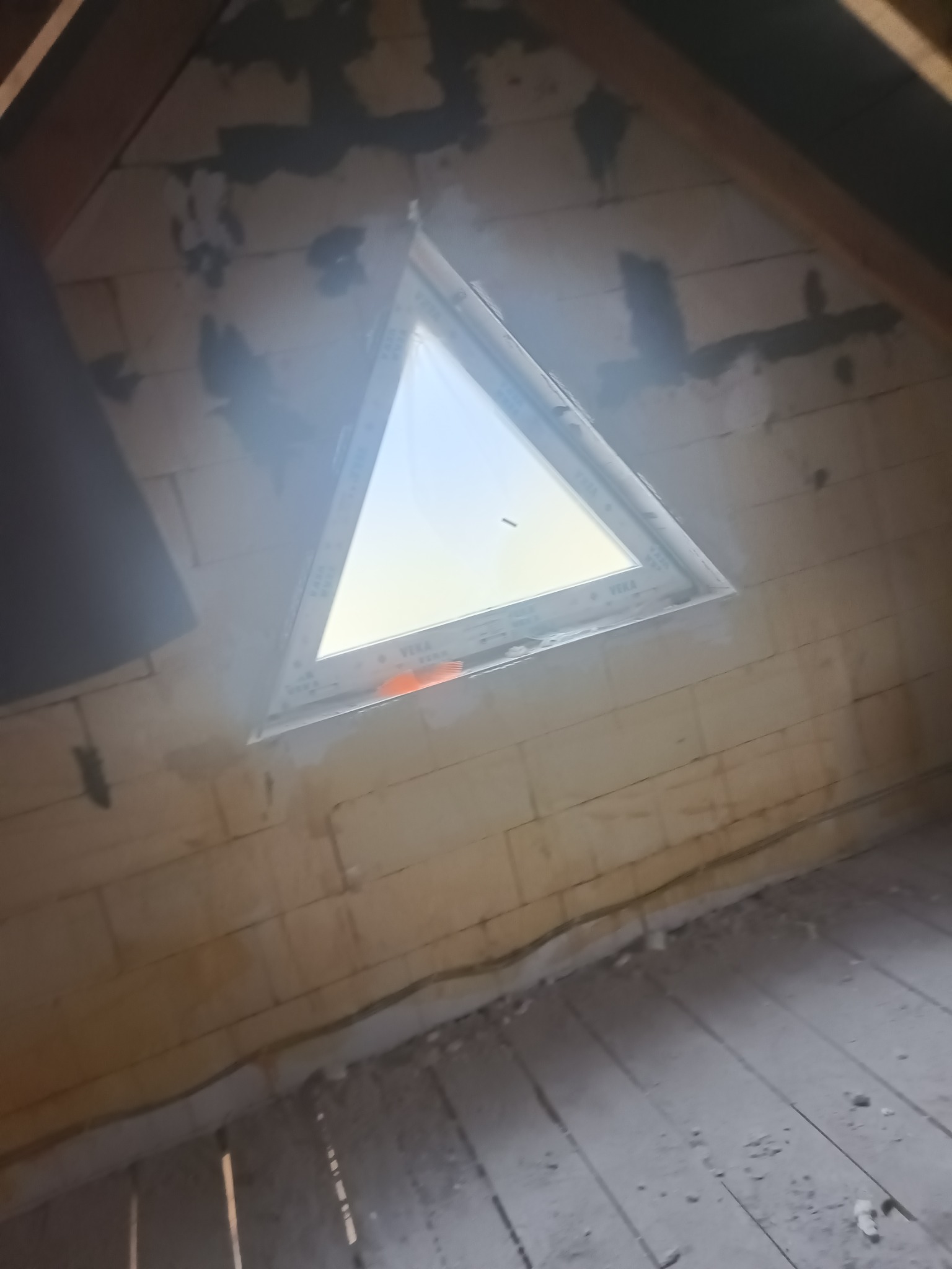 Poddasze z trójkątnym oknem, ściany z pustaków, częściowo przygotowane pod tynkowanie. Widoczne ślady po łatach tynku. Podłoga drewniana, surowa.