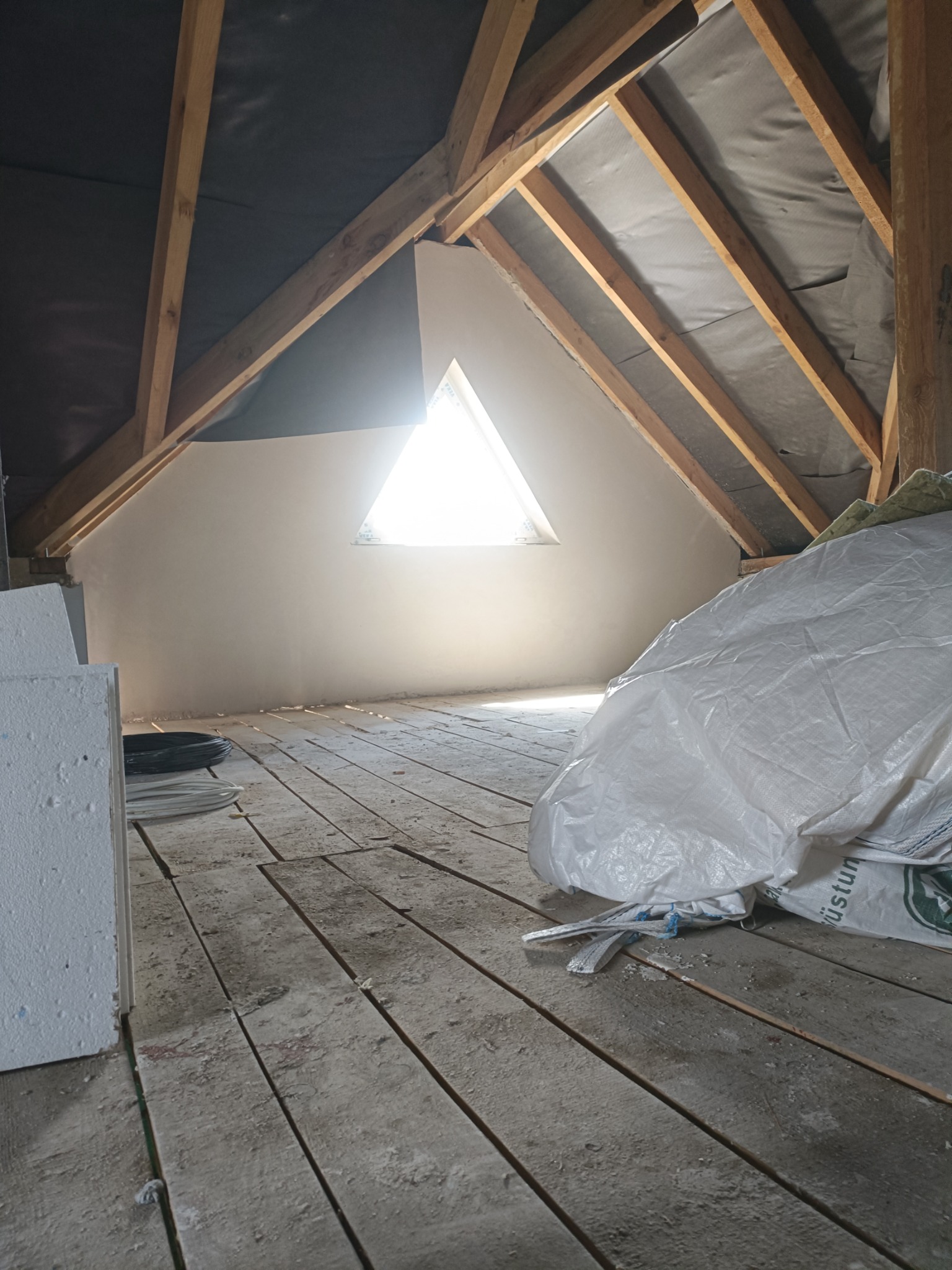 Poddasze z oknem w kształcie trójkąta po nałożeniu tynków. Widoczne drewniane elementy konstrukcyjne dachu i worki z materiałami budowlanymi na podłodze.