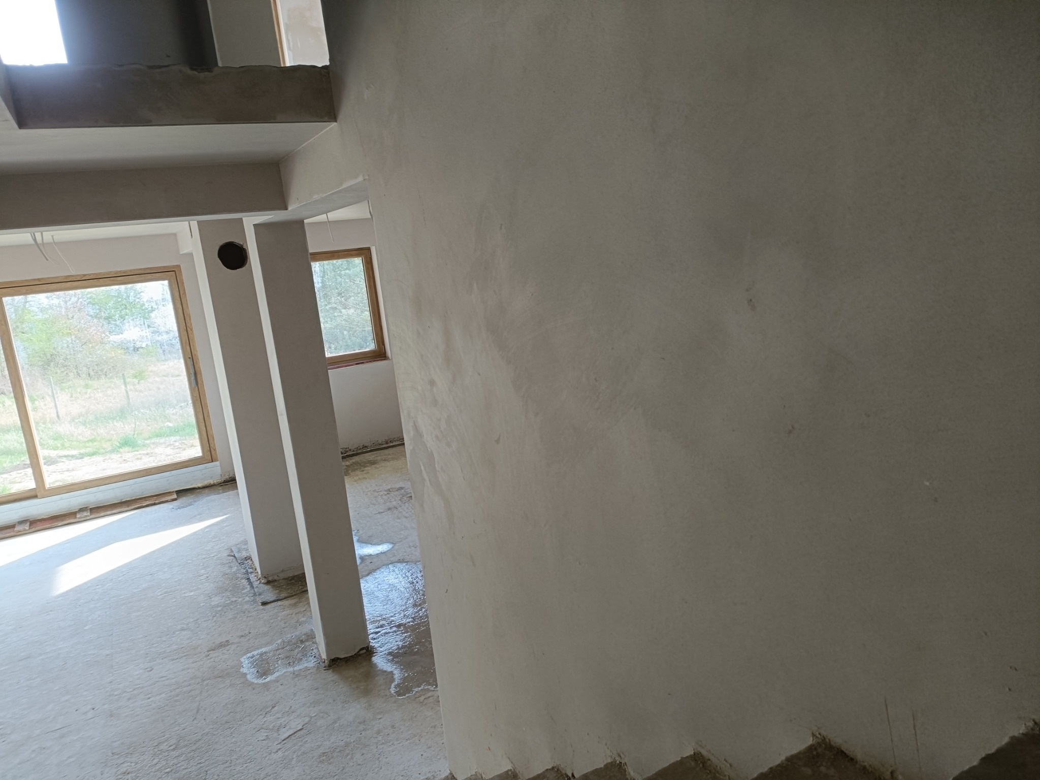 Ściana po nałożeniu tynku maszynowego w surowym wnętrzu budynku z oknami. Widoczne ślady wody na posadzce i kolumny konstrukcyjne.