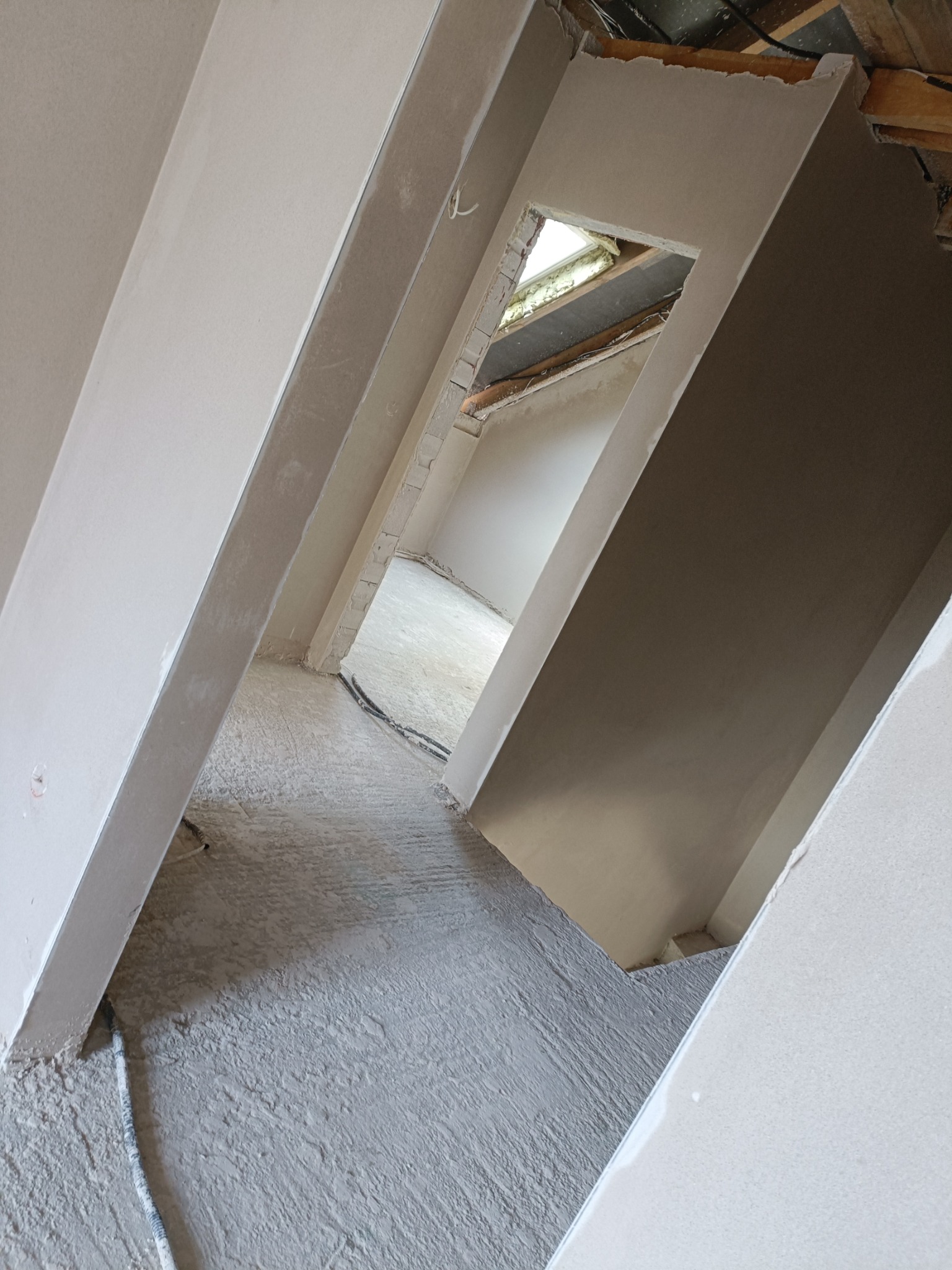 Wnętrze po tynkowaniu maszynowym: świeżo otynkowane ściany i podłoga, widoczne kable, surowy stan wykończenia z oknem w dachu w tle, perspektywa z dołu.