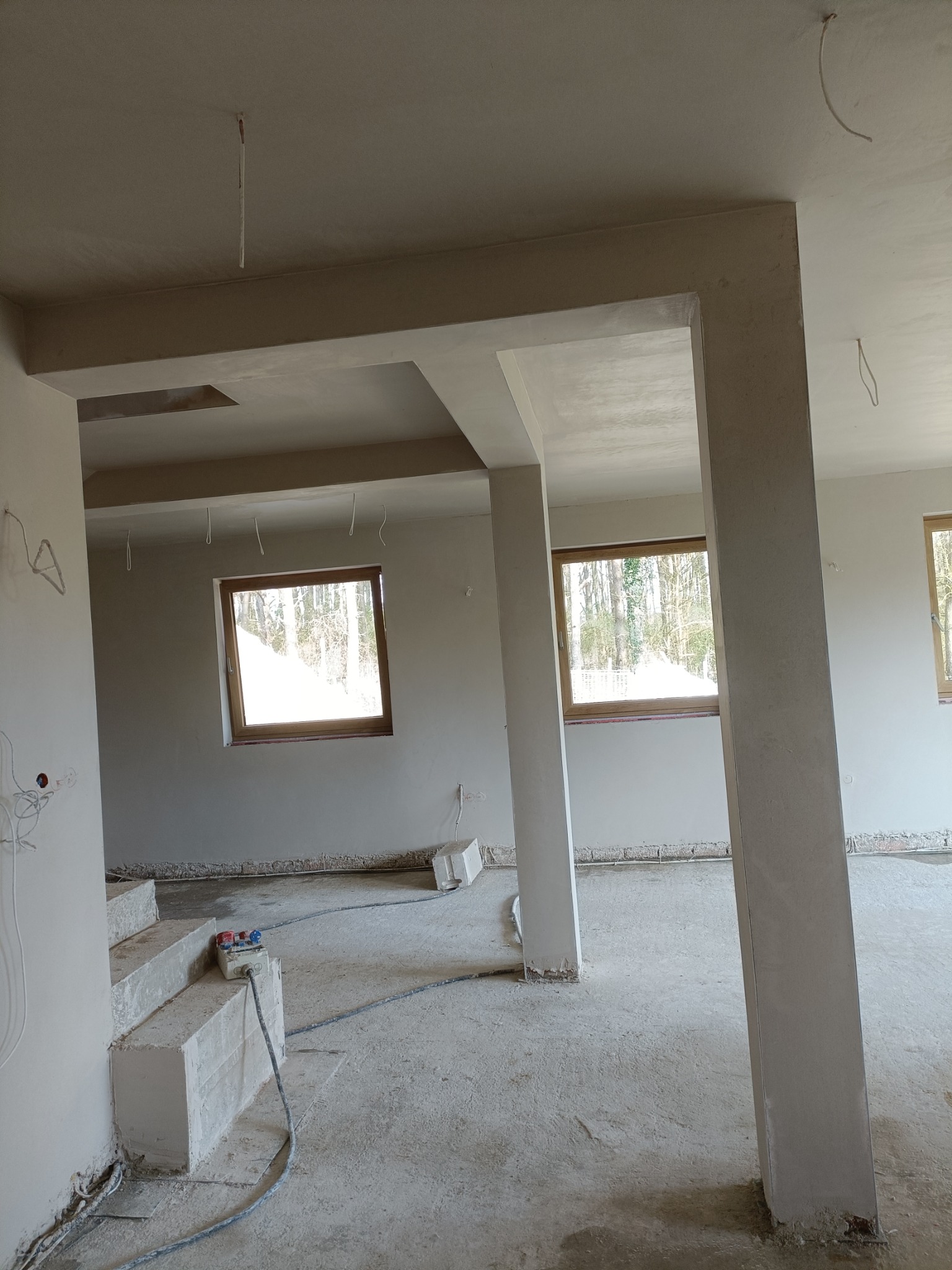 Wnętrze po pracach murarskich: ściany i filary pokryte świeżym tynkiem, okna z drewnianymi ramami, widoczne przewody elektryczne, surowy beton na podłodze.