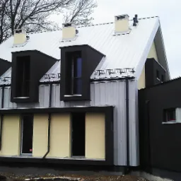 Nowoczesny dom z blachy trapezowej w kolorze srebrnym i czarnymi elementami wykończeniowymi, widoczne trzy lukarny z balkonami i fragment ogrodu.