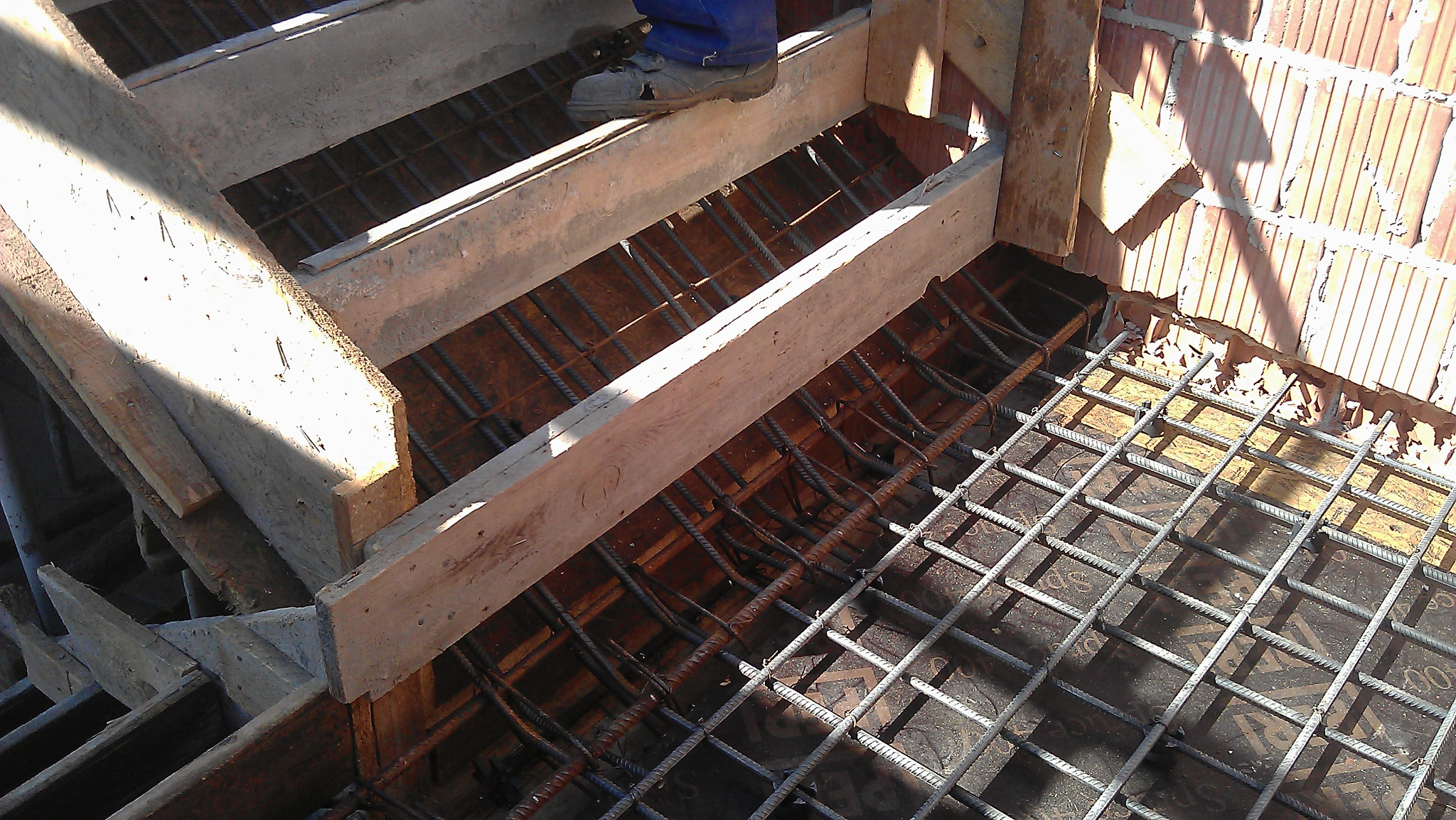 Budowa schodów żelbetowych z drewnianym szalunkiem i siatką zbrojeniową, widoczny fragment muru z cegły dziurawki i noga robotnika w niebieskich spodniach.