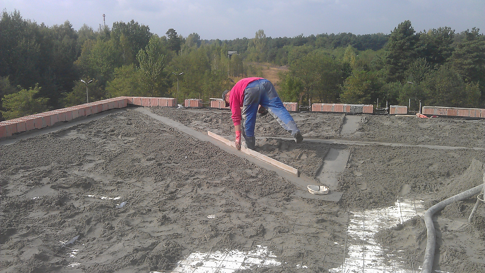 Pracownik w różowej bluzie wyrównuje wylewkę betonową na dachu budynku z użyciem poziomicy, w tle widoczny las.