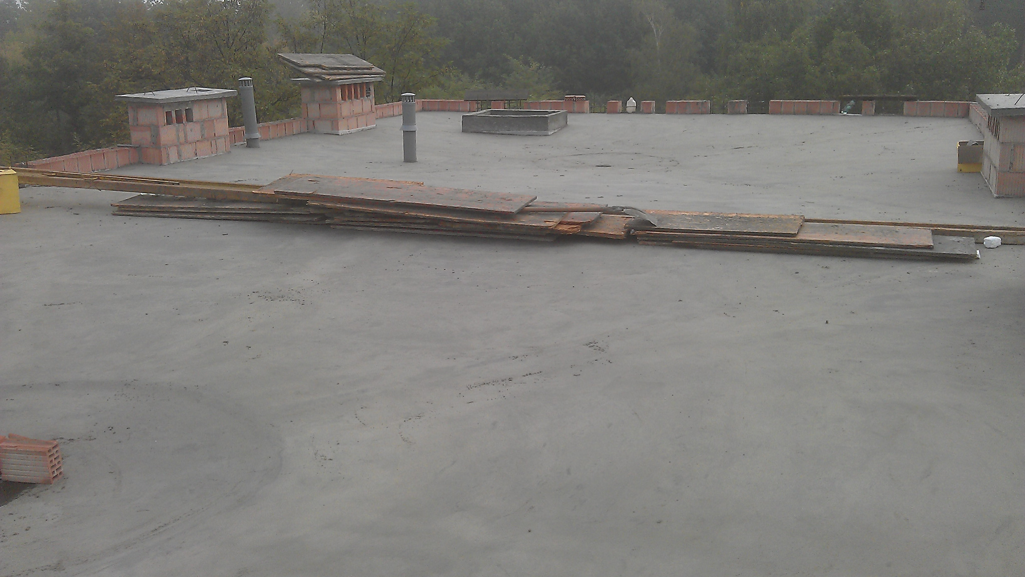 Surowy betonowy dach płaski w trakcie budowy z ceglanymi kominami wentylacyjnymi i stertą starych desek leżących na powierzchni.