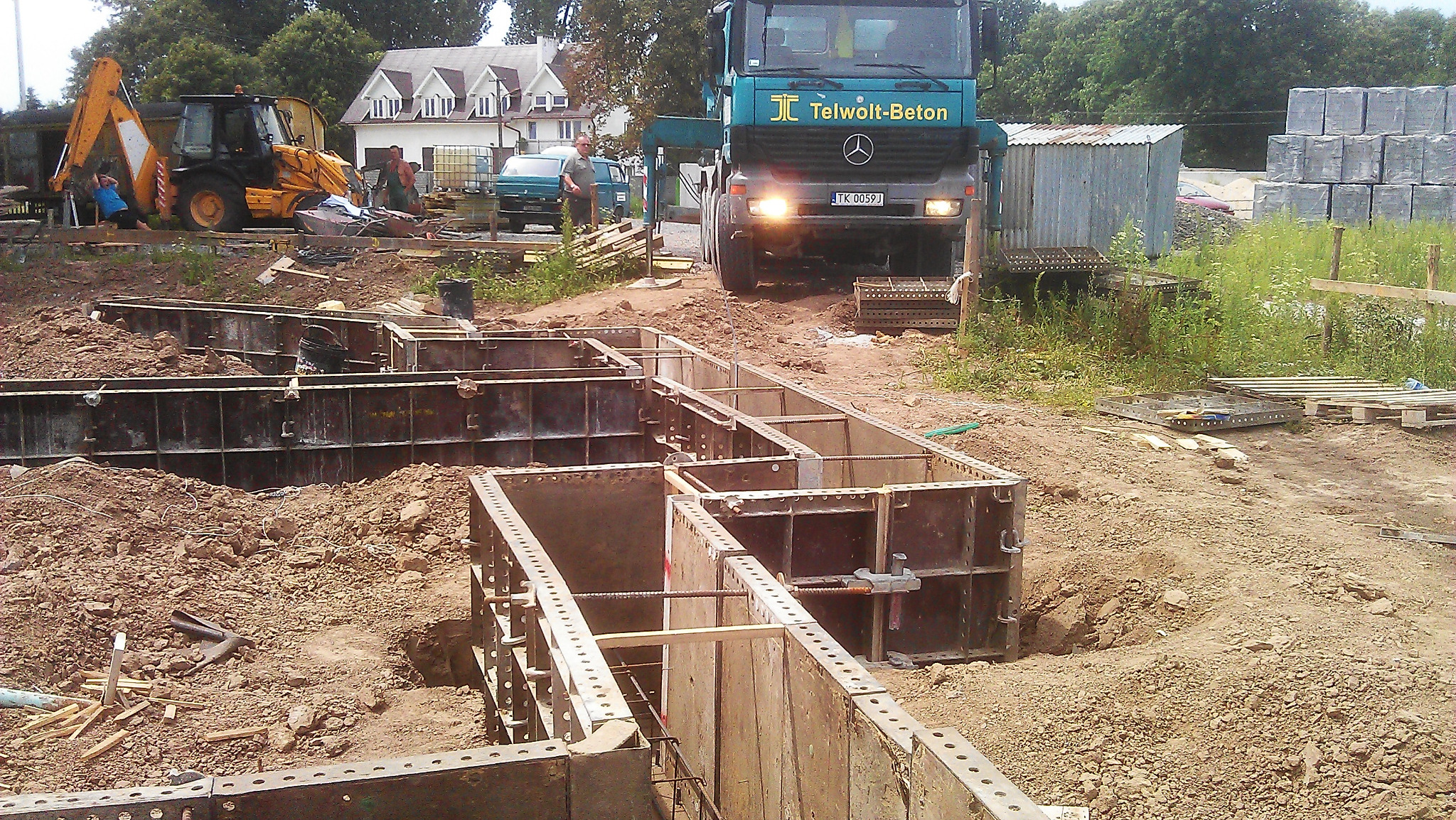 Przygotowanie fundamentów pod budowę domu: szalunki zbrojeniowe, ciężarówka z betonem Telwolt-Beton i koparka w tle.