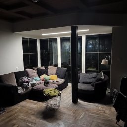 Stanley - Salon z ciemną podłogą w jodełkę, dużym narożnikiem z poduszkami i kotem, oraz ścianą okien z widokiem na nocny ogród.