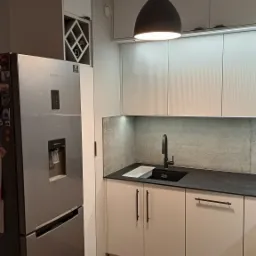 Nowoczesna kuchnia z białymi szafkami, czarnym blatem i zlewem, srebrną lodówką Samsung z dystrybutorem wody i kostkarką, oraz lampą wiszącą z miedzianymi akcentami.