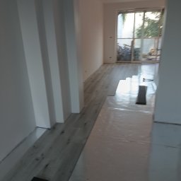 Stanley - Układanie paneli podłogowych w jasnym, odnawianym wnętrzu z widocznym fragmentem okna balkonowego i przygotowanymi panelami.