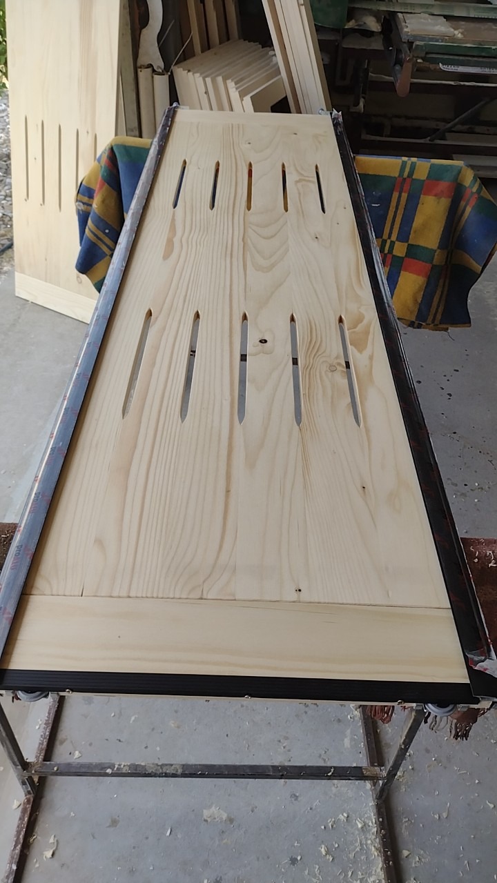 Surowe drewniane drzwi szafy z podłużnymi otworami, oparte na metalowym stojaku, w warsztatowym otoczeniu z widocznymi elementami mebli w tle.