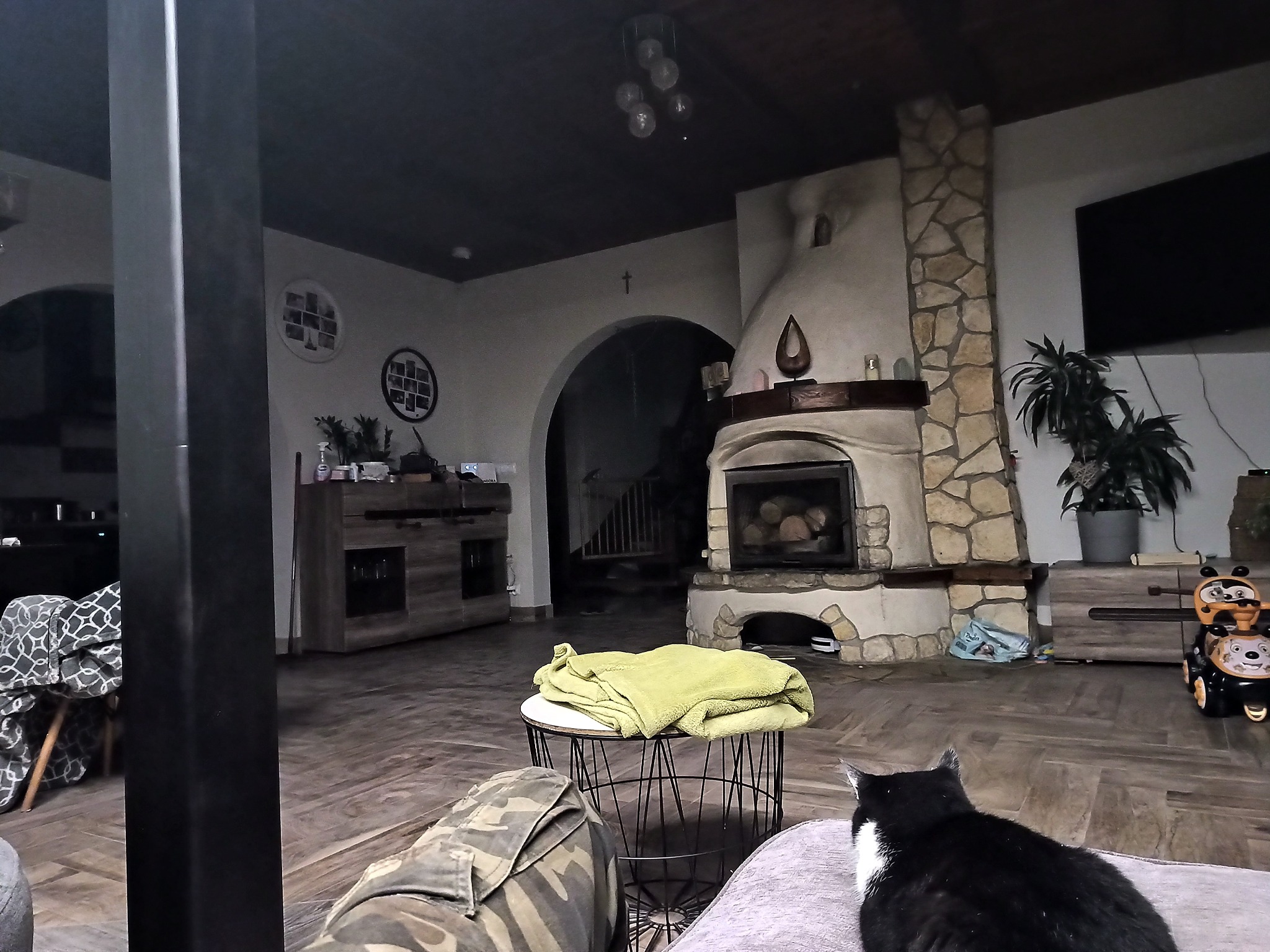 Salon z kamiennym kominkiem i czarno-białym kotem leżącym na kanapie w tle, widok z perspektywy siedzącego na kanapie, słabe oświetlenie.
