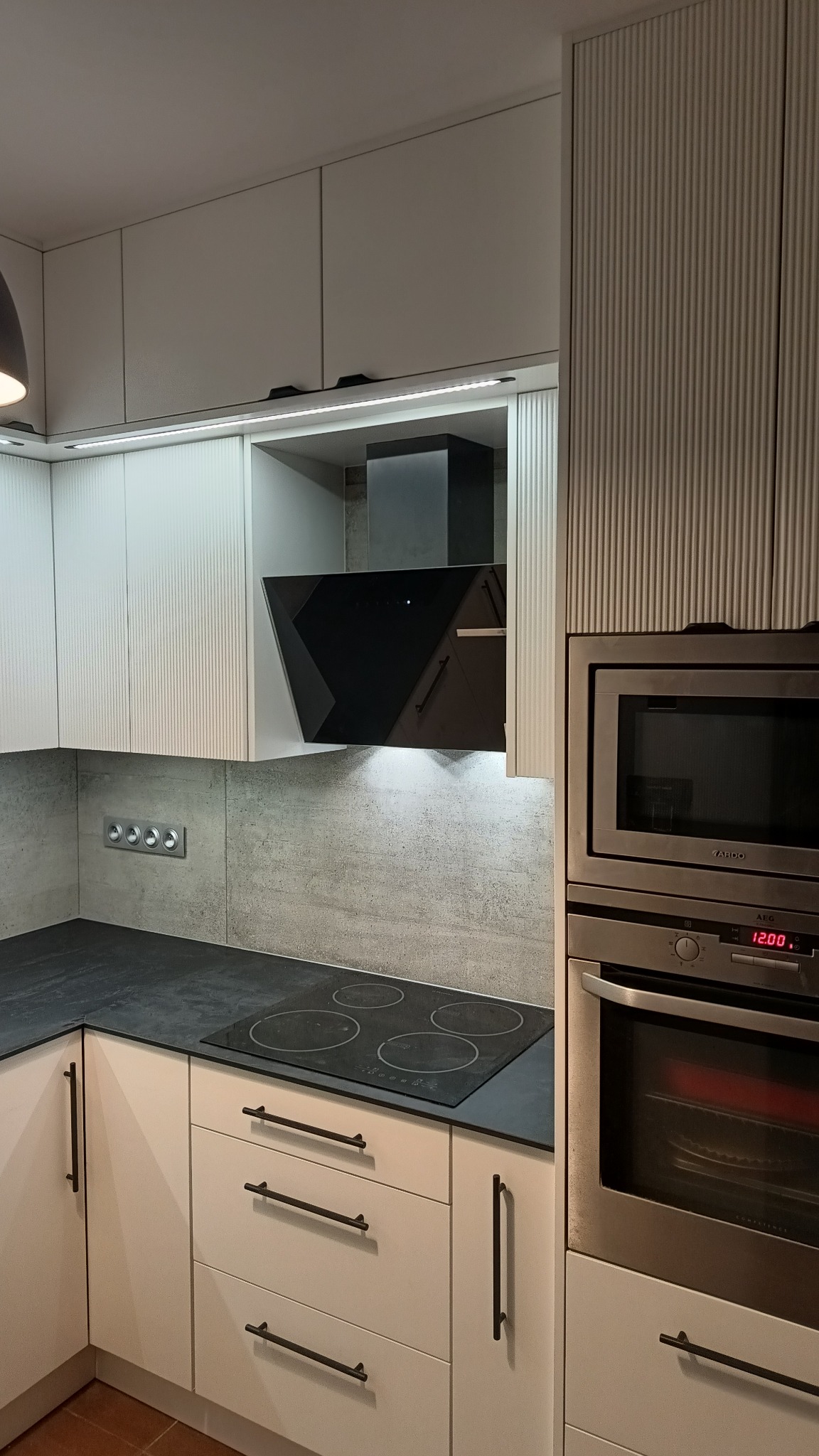 Nowoczesna kuchnia z białymi, ryflowanymi frontami szafek, czarnym blatem, płytą indukcyjną i pochłaniaczem w kolorze czarnym, oświetlenie LED pod szafkami wiszącymi.