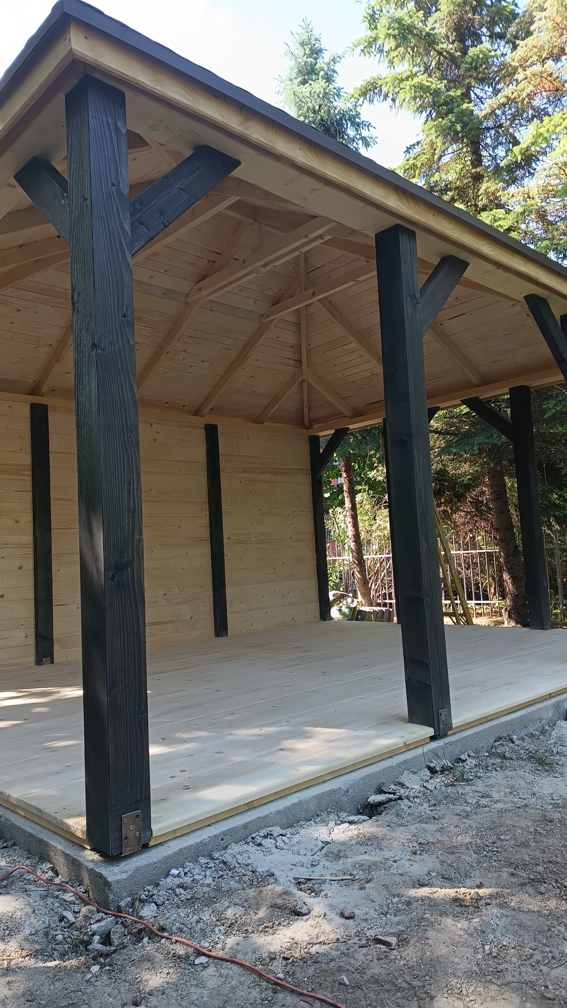 Drewniana altana ogrodowa z ciemnymi, lakierowanymi słupami nośnymi, jasne drewno ścian i podłogi, widoczne elementy konstrukcyjne dachu, w tle zieleń drzew.