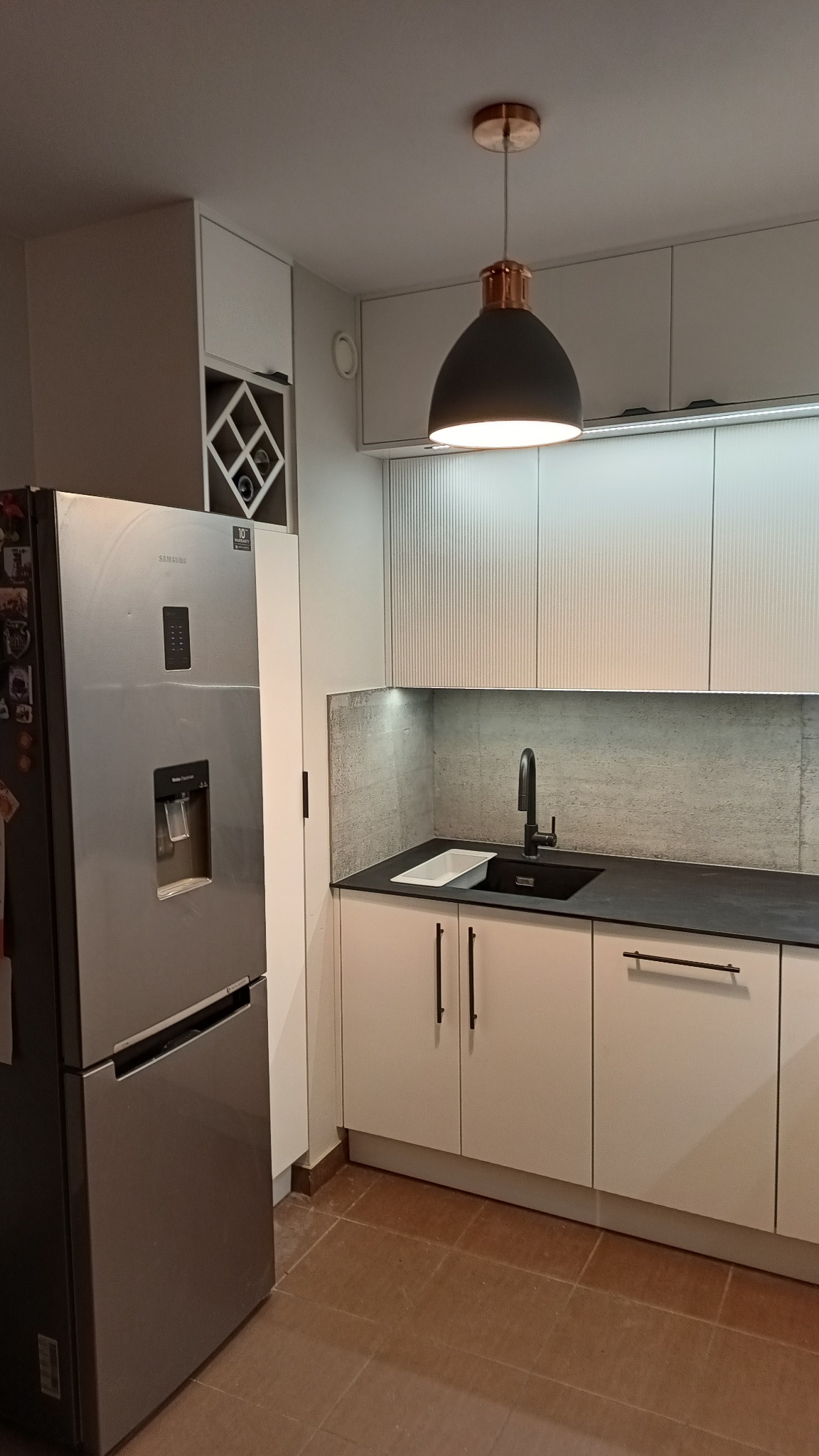 Nowoczesna kuchnia z białymi szafkami, czarnym blatem i zlewem, srebrną lodówką Samsung z dystrybutorem wody i kostkarką, oraz lampą wiszącą z miedzianymi akcentami.