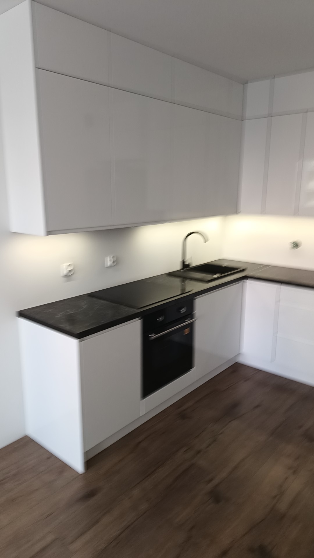 Biała, nowoczesna zabudowa kuchenna z czarnym blatem, wbudowanym piekarnikiem i zlewozmywakiem, oświetlona punktowymi lampkami podszafkowymi.