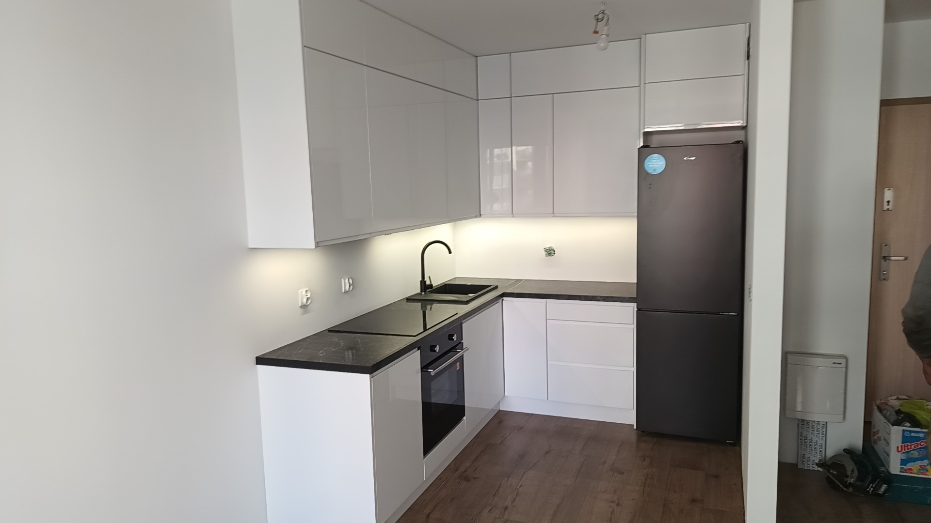 Nowoczesna kuchnia z białymi, lakierowanymi szafkami, czarnym blatem i armaturą, drewnopodobną podłogą i czarną lodówką Candy